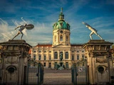 Udsmykkede porte og statuer indrammer det store Charlottenburg-palads i Berlin med en grøn kuppel og et springvand i gården under en blå himmel.