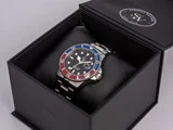 Et sølvfarvet armbåndsur med en blå og rød ramme ligger på en sort pude i en åben sort æske med Schmidt Watches-logoet på låget.