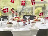Et festligt bord pyntet med danske flag, legetøjssoldater, kager, bær, glas og tallerkener, dækket ved store vinduer med udsigt til haven udenfor.
