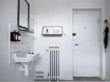 Et hvidt badeværelse med en væghængt håndvask med sæbedispensere, en hylde med små dekorationsgenstande, en radiator og en massiv hvid dør, hvor der hænger en sort paraply i nærheden.