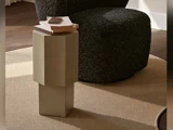 Et lille, moderne beige sidebord med bøger på står på et lysebrunt tæppe ved siden af en mørk stol med struktur i et blødt oplyst rum.