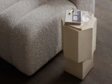 En grå sofa med struktur ved siden af et moderne beige sidebord, hvor der står en bog, en blyant og et glas vand på et mørkt trægulv.