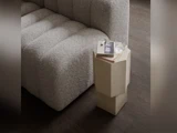 En grå sofa med struktur står ved siden af et beige, geometrisk sidebord, hvorpå der ligger en lukket bog, et par briller og et glas vand på et mørkt trægulv.