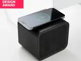 En sort trådløs højttaler med en smartphone til opladning på toppen, vist under en Design Award-label i øverste venstre hjørne.