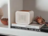 En lille hvid højttaler står på en hylde oven på to bøger med titlerne "ernesto neto" og "CONSULTING THE BOOK" ved siden af en tørret plante og en dekorativ skål.