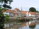 En grøn og rød træbåd, der ligger fortøjet i en kanal foran en række traditionelle europæiske huse med orange tegltage.