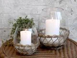 To tændte hvide lys i glasholdere med flettede kurvefødder er placeret på en kurvebakke med en grøn plante og en struktureret grå væg i baggrunden.