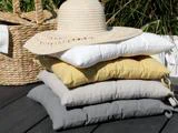 En stak med fire puder i hvid, gul, beige og grå, toppet med en stråhat. En flettet kurv og højt græs står i baggrunden på en træplade.