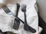 Fire sorte metalgafler er anbragt på en lys linnedserviet, hvor håndtagene og tænderne overlapper hinanden.