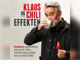 En mand med gråt hår og skæg holder et glas mælk og en brændende chilipeber foran en dansk plakat, hvor der står Klaus og Chili Effekten.