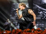 En mandlig kunstner med tatoveringer synger lidenskabeligt på scenen, mens han knæler ved siden af orange blomster iført en lædervest med nitter; en bandkammerat spiller guitar i baggrunden.