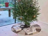 Et pyntet juletræ med et par indpakkede gaver under, placeret på et vævet tæppe i et rum med trægulv og et stort vindue.