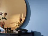 Et rundt spejl på en blå væg reflekterer blomster i en vase og en plante, mens sollyset kaster skygger. På et mørkt bord nedenunder ligger plejeartikler og en rektangulær kasse.