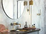 Et moderne badeværelse med marmorvæg, rundt spejl, guldarmaturer, en bordplade med toiletartikler og en væghængt lampe ved siden af et bruseområde.