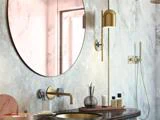 Moderne badeværelse med et rundt vægspejl, marmorvægge, vandhane i guld, bordplade med toiletartikler og en hængende lysarmatur i guld. Et bruseområde er synligt i baggrunden.