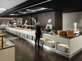 Et moderne cafeteria med kokke i hvide uniformer, der serverer mad bag en disk, mens en kvinde i forretningstøj taler med en af dem. Tallerkener og kopper er pænt stablet under disken.