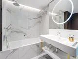 Moderne badeværelse med hvide marmorvægge, badekar med bruseafskærmning i glas, dobbelt håndvask og et rundt, oplyst spejl over håndvasken. Håndklæder og toiletartikler er pænt arrangeret.