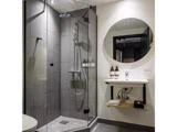 Moderne badeværelse med en glasafskærmet bruser, grå fliser, et rundt spejl over en hvid håndvask og toiletartikler på både brusevæggen og håndvaskhylden.