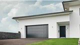 Moderne hvidt hus med en stor grå garageport, sort hoveddør, skrånende indkørsel og grøn græsplæne under en overskyet himmel.