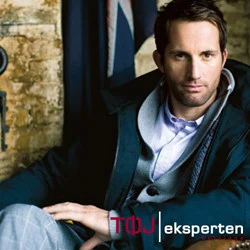 En mand i en mørk frakke sidder indendørs og kigger ind i kameraet. Bag ham ses en del af et flag og en struktureret væg. Nederst står teksten TØJ eksperten.