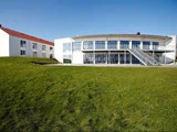 En moderne hvid bygning med store vinduer og en udvendig trappe står på en græsklædt bakke under en klar blå himmel. En anden hvid bygning er synlig til venstre.