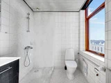 Et lyst badeværelse med bruseniche, hvide fliser, toilet, stort vindue og gråt gulv. Blå himmel og hustage er synlige udenfor.