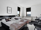 Lyst hotelværelse med en dobbeltseng, to puder, foldede håndklæder, en bænk ved vinduet, to stole, vægkunst og to store vinduer med sorte persienner.