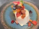 En anrettet dessert består af en cylinder af cremet hvid mousse toppet med rabarber i tern, ved siden af en lille tærte pyntet med blåbær og et dekorativt stykke chokolade på en blå keramiktallerken.