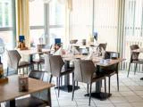 Lys, moderne restaurant med pænt dækkede træborde, grå stole, glas og hvide servietter; store vinduer lukker naturligt lys ind.