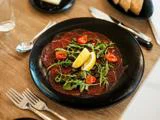 En sort tallerken med tynde skiver rå oksecarpaccio, toppet med rucola, cherrytomater og to citronbåde, på et træbord med bestik og brød i nærheden.