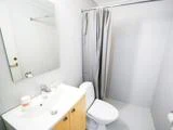 Et lille, rent badeværelse med hvide flisevægge, en håndvask med et spejl over, et toilet og et bruseområde med et gråt forhæng. Der står toiletartikler på håndvasken.