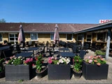 Udendørs terrasse med sorte borde og stole, lyserøde og hvide blomster i potter foran, store parasoller og en murstensbygning i baggrunden under en klar blå himmel.