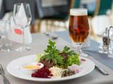 En tallerken med bøf, grønt, grøntsager i tern, kapers og sauce ved siden af et glas øl på et dækket spisebord med tomme glas i baggrunden.