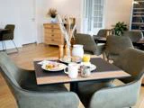 Et morgenbord med brød, kødskiver, ost, salat, appelsinjuice, en kande, kaffekrus, salt- og peberbøsser og en sukkerskål i en hyggelig spisestue med grønne stole.