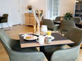 Morgenbord dækket til to med tallerkener med mad, krus, appelsinjuice, brød, salt- og peberbøsse og en hvid kande i en hyggelig, moderne spisestue med grønne stole.