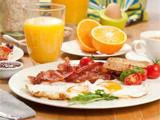 En morgenmadstallerken med spejlæg, bacon, ristet brød, cherrytomater og grønt ved siden af et glas appelsinjuice, appelsinskiver, et æg og en kop kaffe på et træbord.