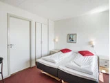 Et simpelt hotelværelse med to enkeltsenge skubbet sammen, hvidt sengetøj, røde puder, natborde, væglamper, et klædeskab og en dør.