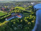 Luftfoto af en stor hvid bygning med rødt tag omgivet af træer nær kysten, med en by og åbne marker i baggrunden.
