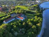 Luftfoto af en stor bygning omgivet af træer nær en kystlinje med huse, marker og en by i baggrunden.