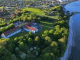 Luftfoto af en stor bygning med rødt tag omgivet af træer nær en kystlinje, med en by og vand i baggrunden.