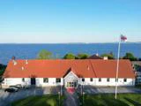 En stor hvid bygning med rødt tag ligger ved en rolig blå sø med en flagstang med et hvidt flag foran og en klar blå himmel over.
