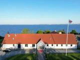 En hvid bygning med rødt tag står ved et stort vandområde med træer bagved og en flagstang med et flag foran. Himlen er klar og blå.
