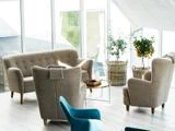 En lys, moderne stue med beige lænestole, et lille træbord, potteplanter i flettede kurve og store vinduer, der lukker naturligt lys ind.