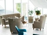 Moderne stue med beige lænestole og sofa, en blå stol, sofabord i glas og potteplanter i flettede kurve ved store vinduer, der lukker naturligt lys ind.