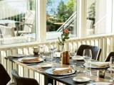 Et solbeskinnet restaurantbord dækket til fire med tallerkener, glas, servietter og en enkelt lyserød rose i en vase nær store vinduer med udsigt til en lys udendørs terrasse.