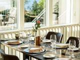 Et solbeskinnet restaurantbord dækket til fire med glas, tallerkener, servietter og en enkelt lyserød rose i en vase ved siden af store vinduer med udsigt til en udendørs terrasse.
