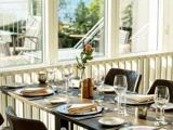 Et solbeskinnet restaurantbord dækket med tallerkener, glas, servietter og en enkelt lyserød rose i en vase ved siden af store vinduer med udsigt til en lys udendørs gårdhave.