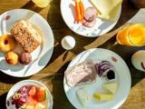 Morgenmaden er dækket på et træbord med tallerkener med brød, kager, ost, pålæg, grøntsager i skiver, frugt, et helt æg og et glas appelsinjuice.