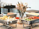 Et buffetbord med forskellige skiveskårne oste, kød og grøntsager, dækket omkring en vase med tørrede blomster, med tallerkener og skåle stablet i baggrunden.