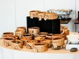 En række mærkede papirposer fyldt med forskellige nødder, frø og tørrede frugter er arrangeret på et bord, sandsynligvis til en morgenmadsbuffet eller snackbar.